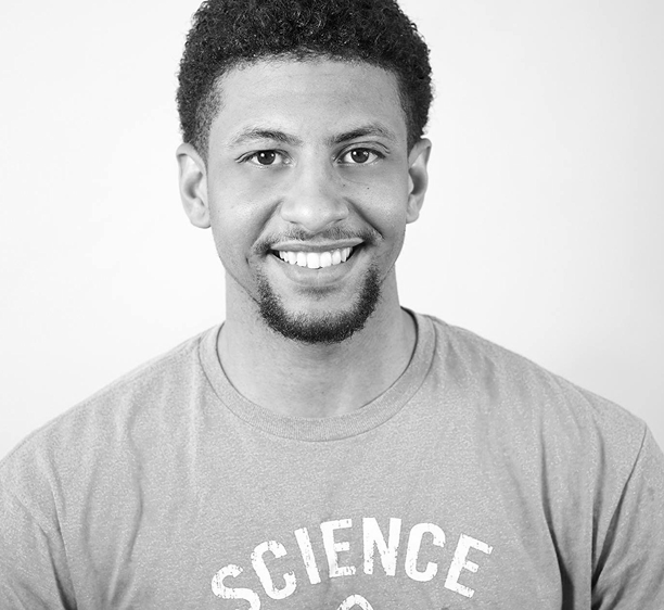 Black-and-white photo of a smiling young man with short curly hair, a trimmed beard, and mustache, wearing a T-shirt that partially reads SCIENCE. He is facing the camera against a plain background.