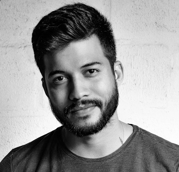 A man with short dark hair and a beard is wearing a crew-neck t-shirt, looking at the camera with a slight smile. The image is black and white, with a textured wall in the background.