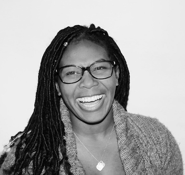Black and white photo of a woman with long braided hair and glasses, wearing a knitted sweater and a heart necklace, smiling widely and looking at the camera.