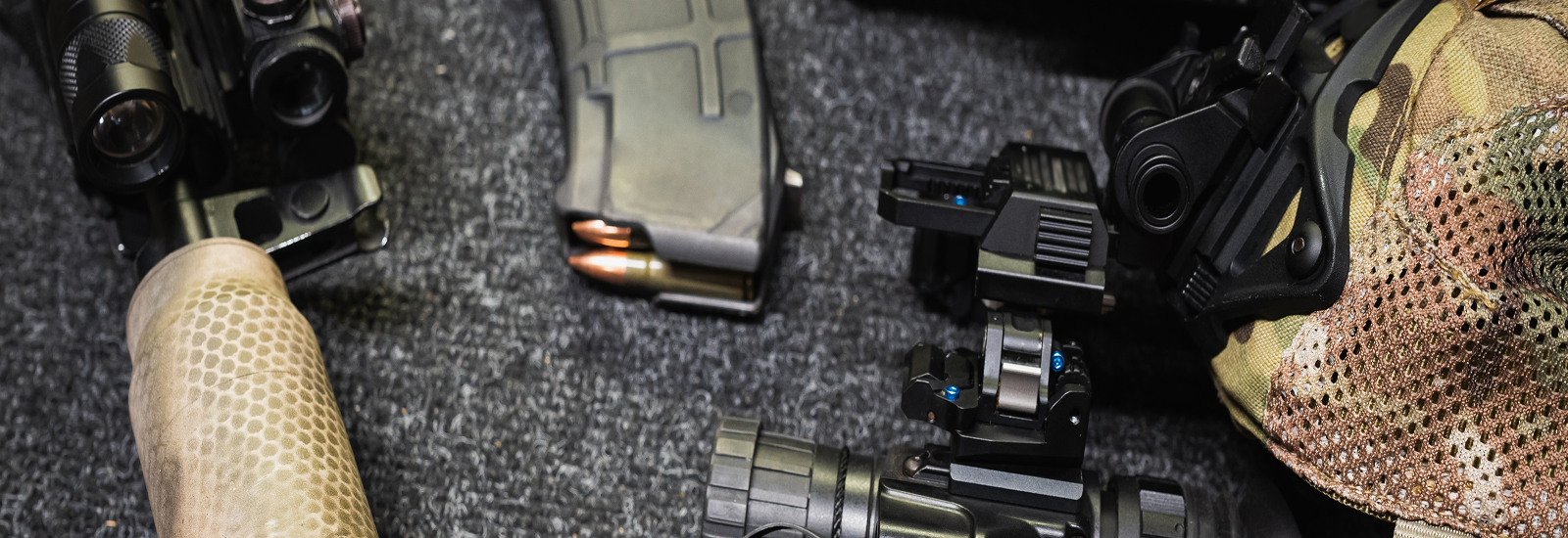 Close-up of tactical gear laid out on a dark textured surface, including a camouflage glove, a magazine with bullets, scopes, and various firearm accessories.