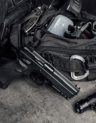 A black handgun, flashlight, folding knife, and various tactical gear are visible partially inside and around an open black backpack on a concrete surface.