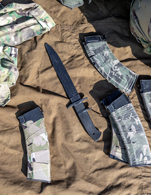 A combat knife and four camouflage-covered rifle magazines are arranged on a brown fabric surface, with military gear partially visible around the edges.